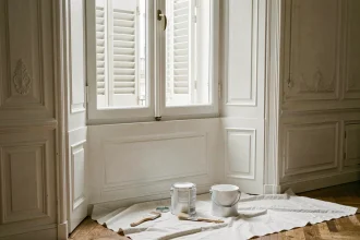 Boiserie de fenêtre en bois peinte en blanc mat dans un appartement haussmannien, avec pot de peinture et pinceaux posés sur une bâche de protection
