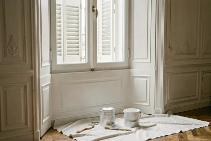Boiserie de fenêtre en bois peinte en blanc mat dans un appartement haussmannien, avec pot de peinture et pinceaux posés sur une bâche de protection